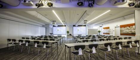 A conference room with rows of tables and chairs, ceiling-mounted projectors, and a large screen.