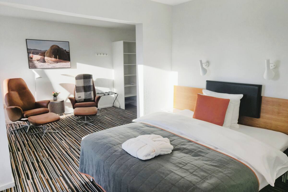 A hotel room with a large bed, a chair, a small table, and a bookshelf.