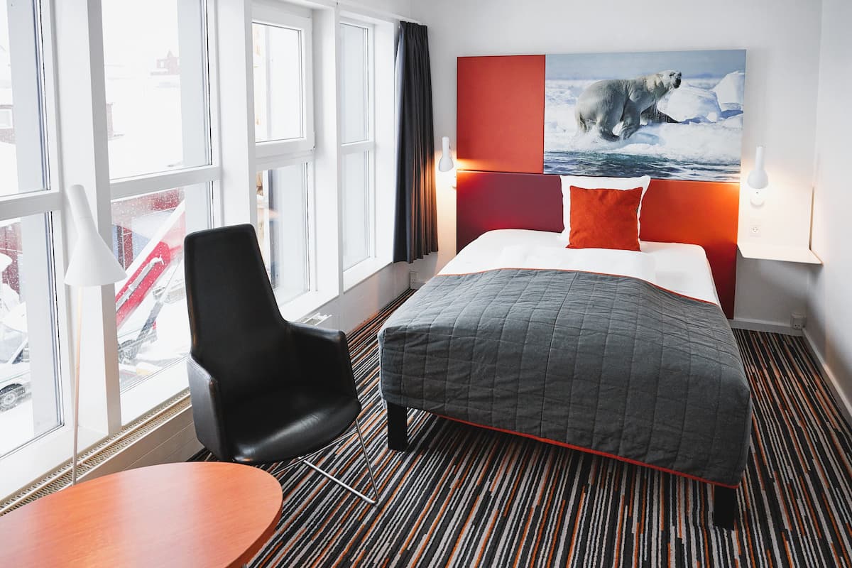 A hotel room with a bed, a chair, a table, a window with a view of a red vehicle outside, and a polar bear painting on the wall.