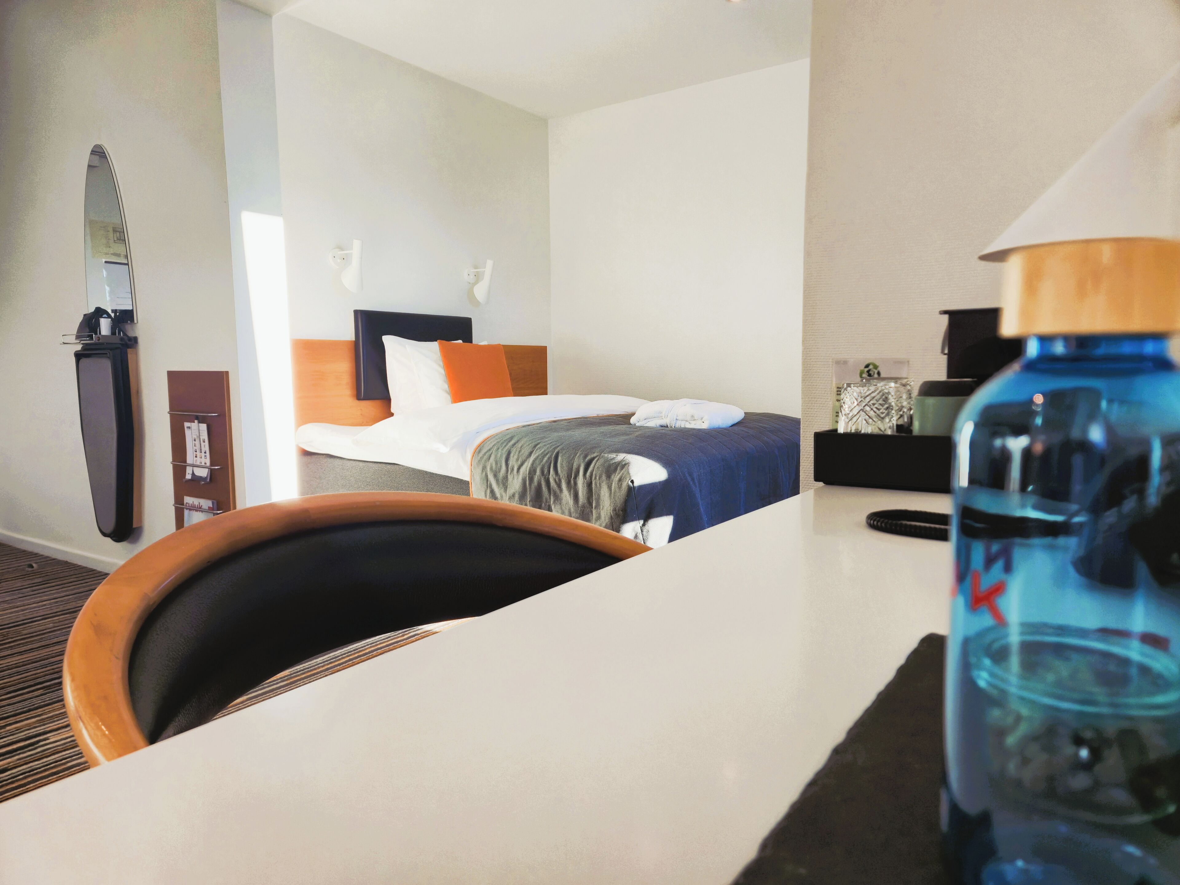 A hotel room with a bed, a desk, a chair, a water bottle, and a mirror.