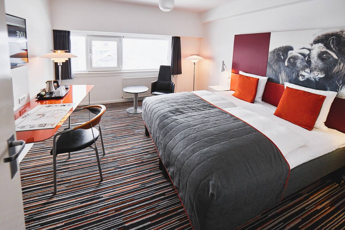 A hotel room with a large bed, a desk with a chair, a small table, and a window.