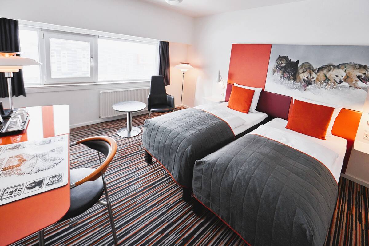 A hotel room with two beds, a desk, a chair, a window with curtains, a floor lamp, and a painting of huskies on the wall.