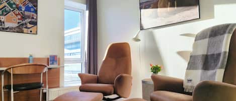 A hotel room with a brown leather armchair, a matching ottoman, a small table with a water bottle, a framed picture of a walrus, and a window with a view of buildings.