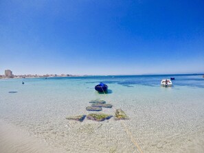A beach with clear shallow waters, boats, and a distant city skyline.