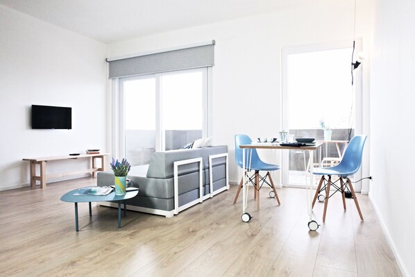 A modern living room with a grey sofa, a small table with a vase, and a dining area with two blue chairs and a table.