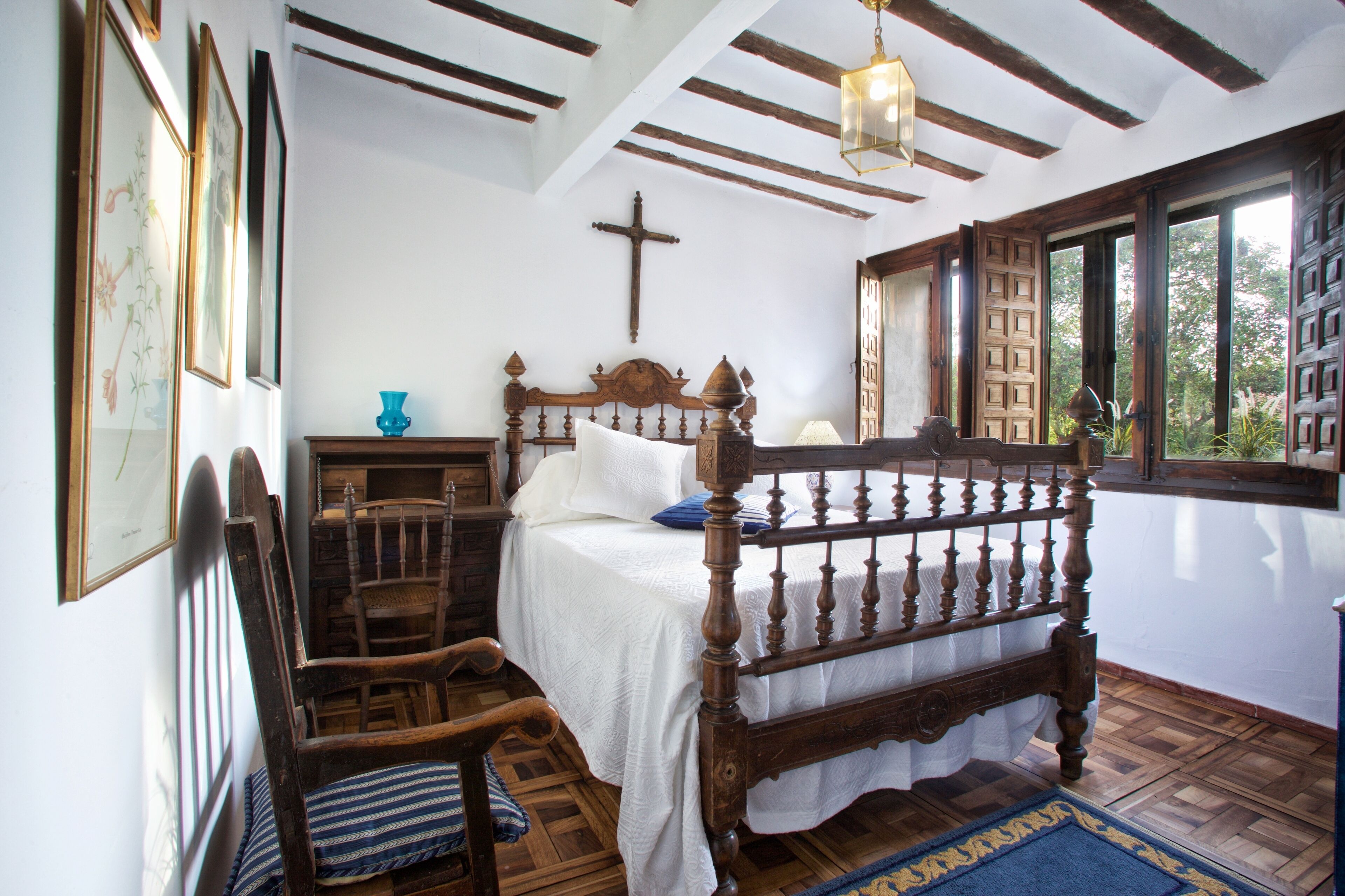 Un dormitorio con una cama de madera, una cruz en la pared y una ventana con vistas a la vegetación.