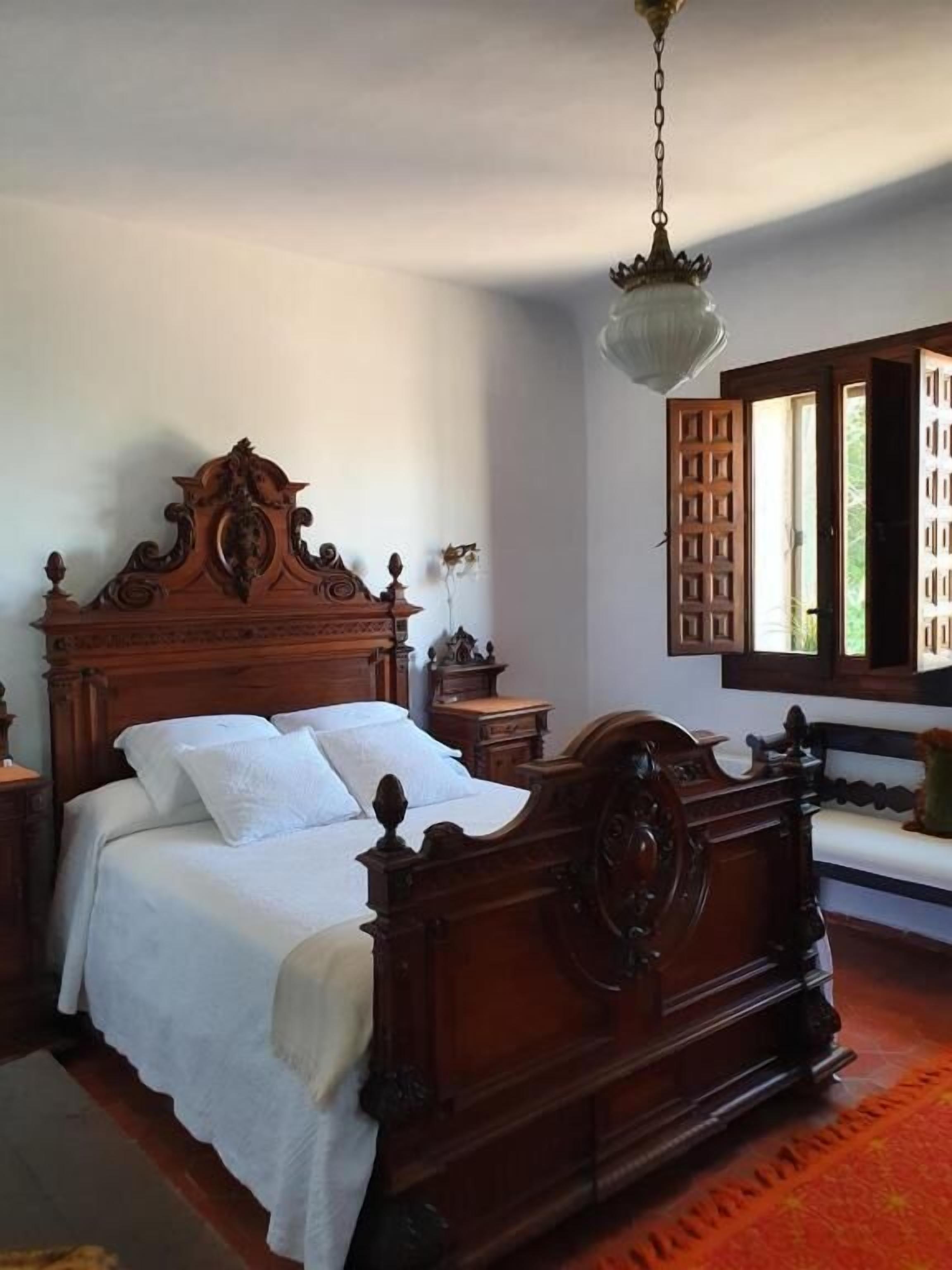 Una cama de madera tradicional con cabecero alto, una mesita de noche y una ventana con contraventanas de madera.