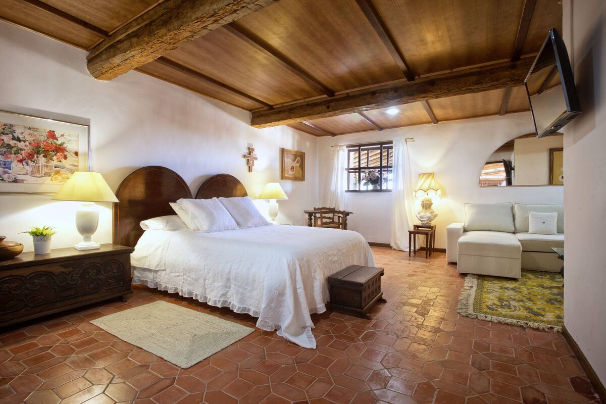 Quarto com cama grande, cabeceira de madeira, luminárias de cabeceira, mesa de cabeceira, uma pequena mesa, um sofá e uma TV fixada na parede.