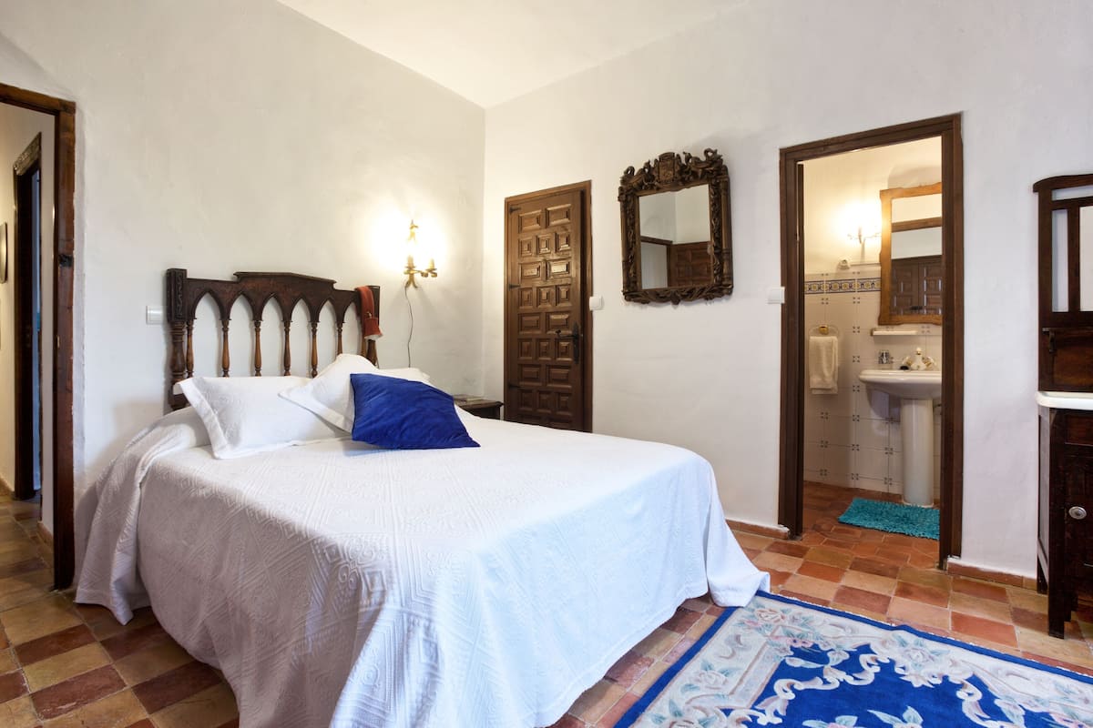 Quarto com cama, cabeceira de madeira, espelho e banheiro visível por uma porta aberta.