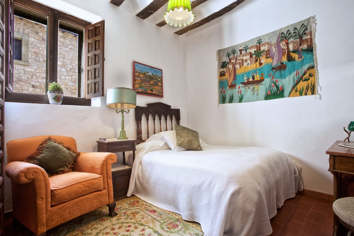 Quarto com cama, poltrona, mesa de cabeceira, luminária, pintura e janela com vista para um edifício.