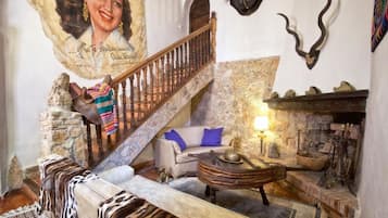 Un interior rústico con una escalera de piedra, un sofá con una manta estampada, una mesa redonda de madera y una chimenea con repisa de piedra.