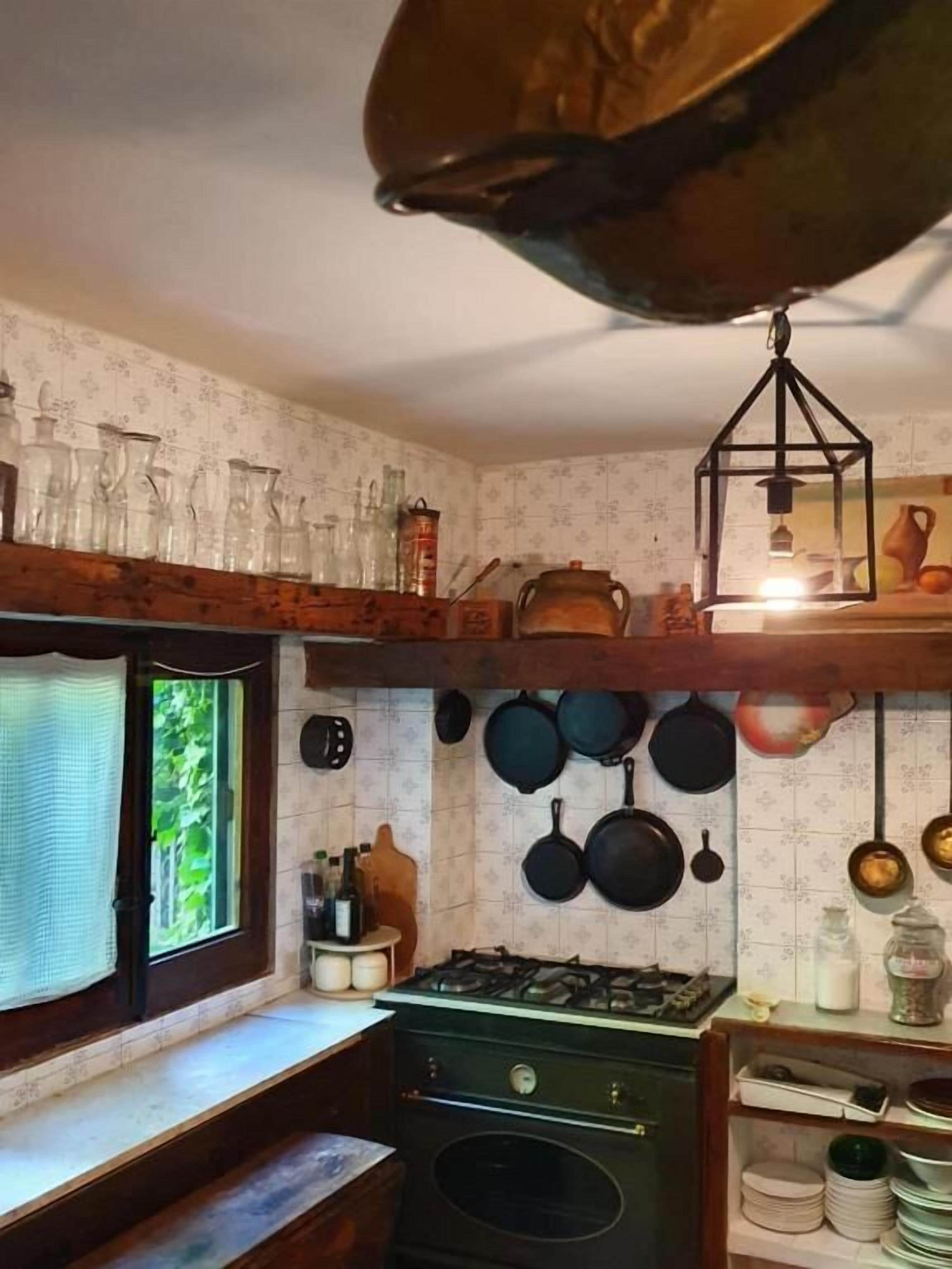 Une cuisine équipée d’une cuisinière à gaz, avec des ustensiles de cuisine suspendus et une fenêtre donnant sur un paysage verdoyant.