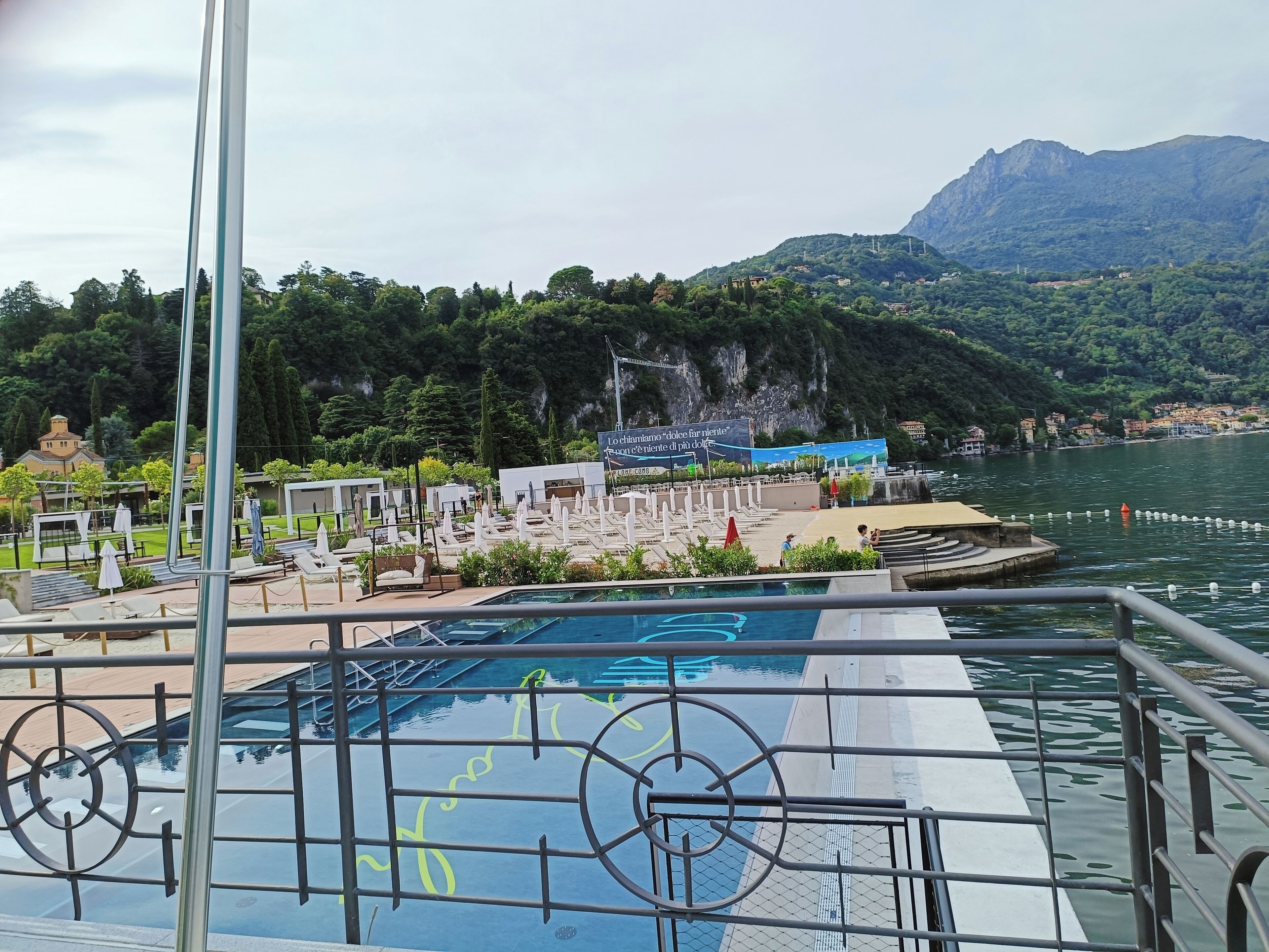 A pool area with lounge chairs and a fountain, surrounded by greenery and overlooking a body of water with mountains in the background.