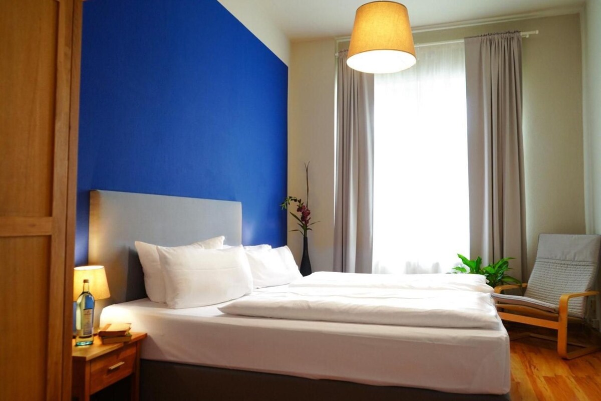 A hotel room with a bed, a nightstand, a lamp, a chair, and a window with curtains.