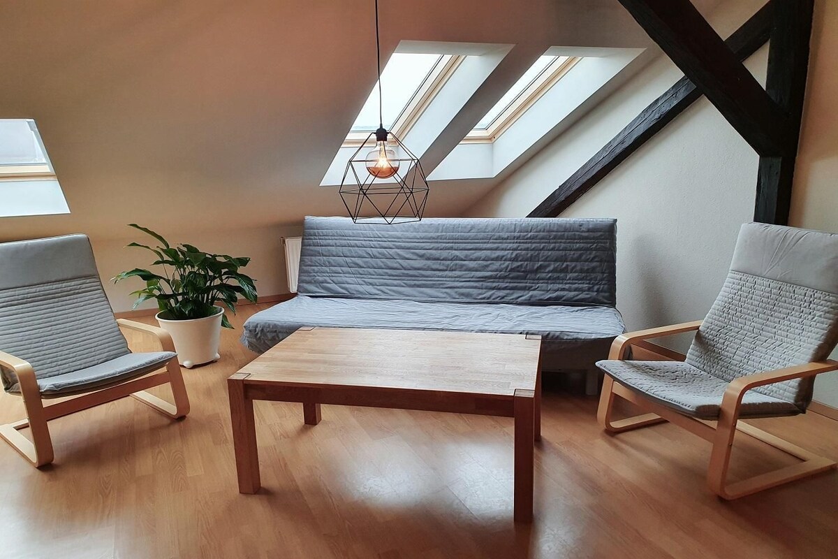 A loft-style room with a wooden floor, a sofa, two armchairs, a hanging light fixture, and a potted plant.