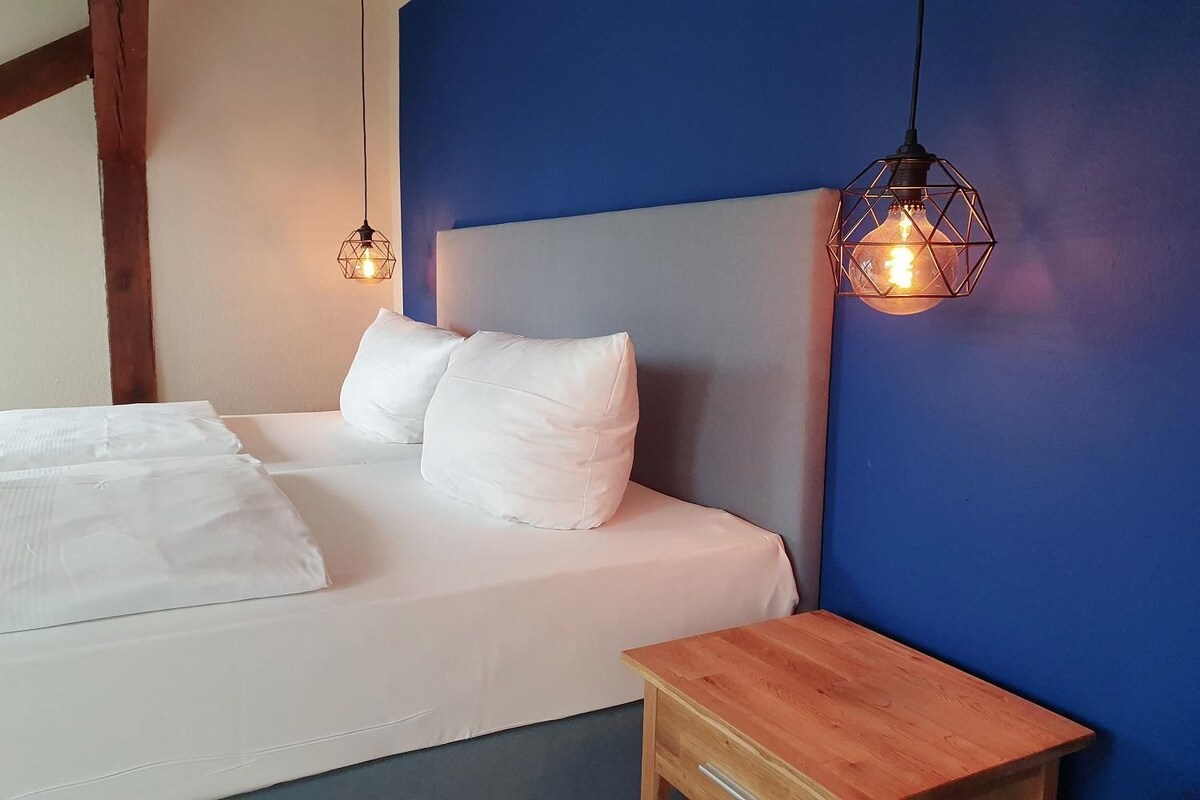 Una cama con sábanas blancas, una mesita de noche de madera y dos lámparas colgantes.