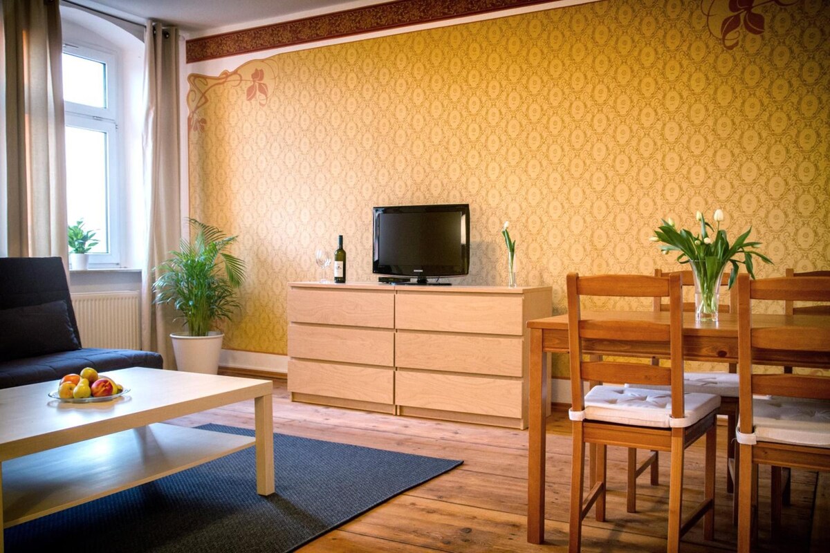 Una sala de estar con una televisión sobre un mueble de madera, una mesa de comedor con sillas, una planta en maceta y una ventana con cortinas.
