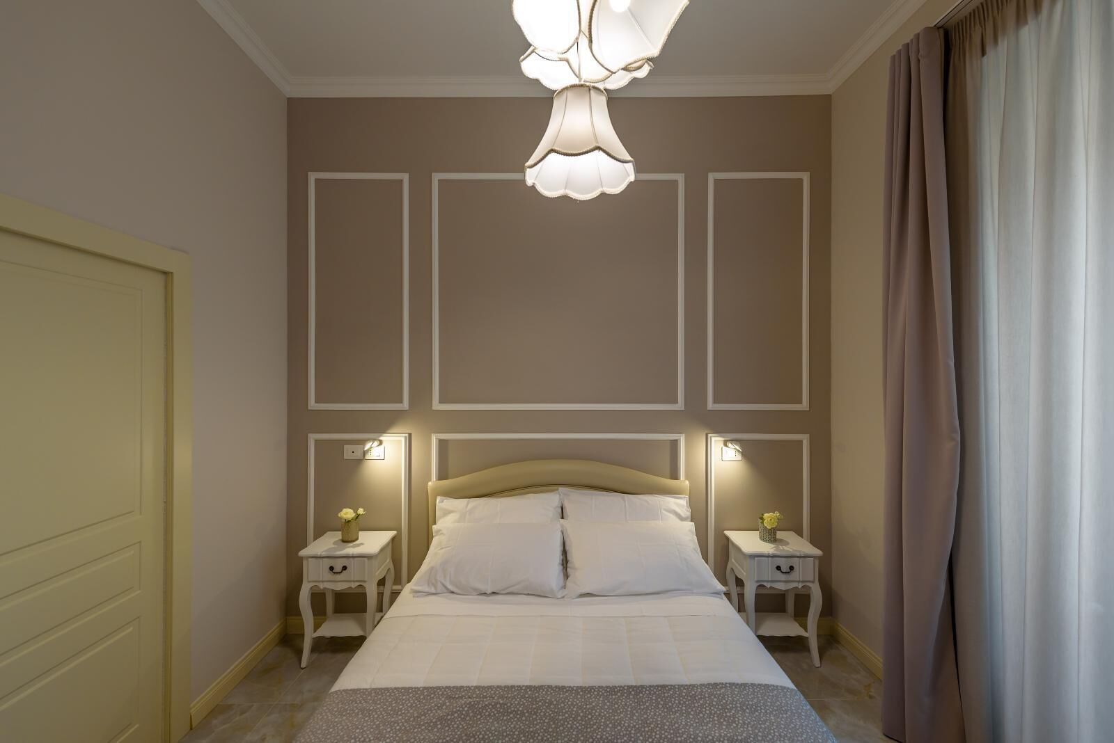 A bedroom with a large bed, two bedside tables with lamps, a decorative ceiling light, and a view of the outside through a window with curtains.