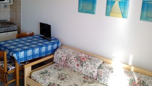 Una habitación compacta con kitchenette, una cama con ropa de cama floral, una mesita con mantel a cuadros azules y dos cuadros en la pared que representan una escena de playa.