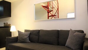 Een moderne woonkamer met een grijze bank, twee gestreepte kussens, een lamp en een rode spiraaltrap op de achtergrond.