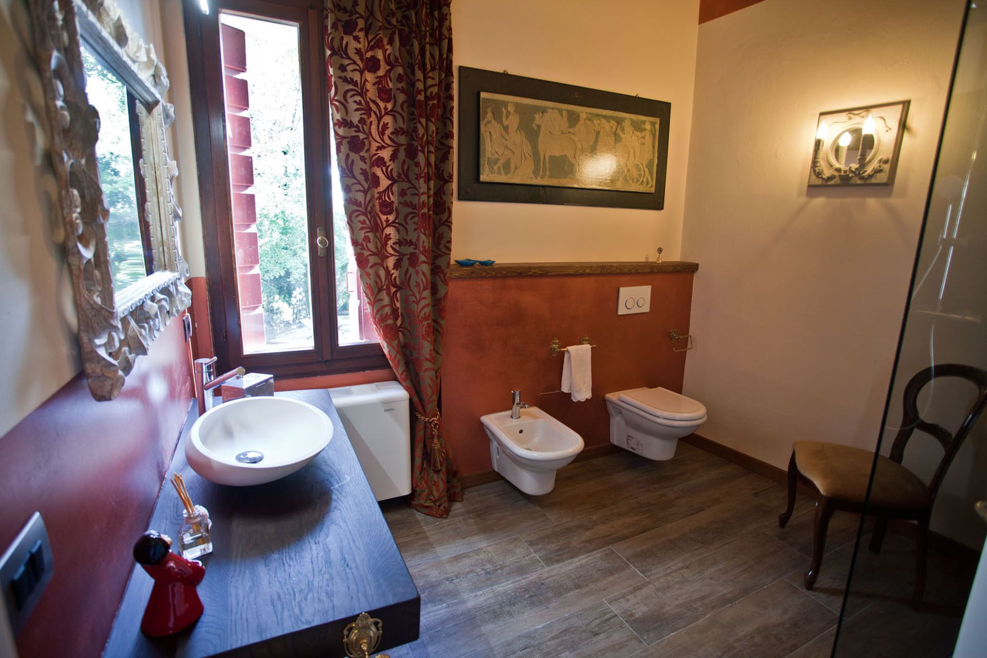 A bathroom with a wooden vanity, a sink, a bidet, and a toilet. There is a window with curtains, a mirror, and a framed picture on the wall.