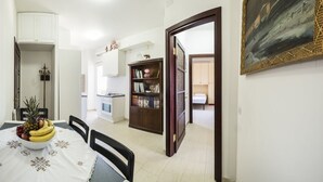A dining area with a table set for two, a kitchen in the background, a bookshelf, and a door leading to another room. - The terrace on the park - bathrooms in the room - panoramic - near the beach (Quartu Sant'Elena)