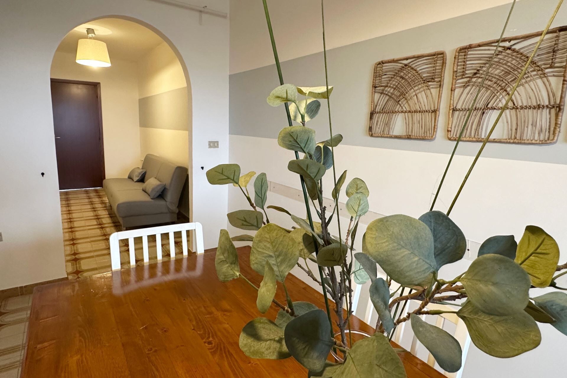 A hallway with a wooden floor, a sofa, and a plant. There are two wall-mounted artworks and a door in the background.