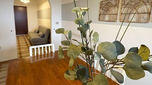 Un pasillo con piso de madera, un sofá y una planta. Se observan dos obras de arte colgadas en la pared y una puerta al fondo.