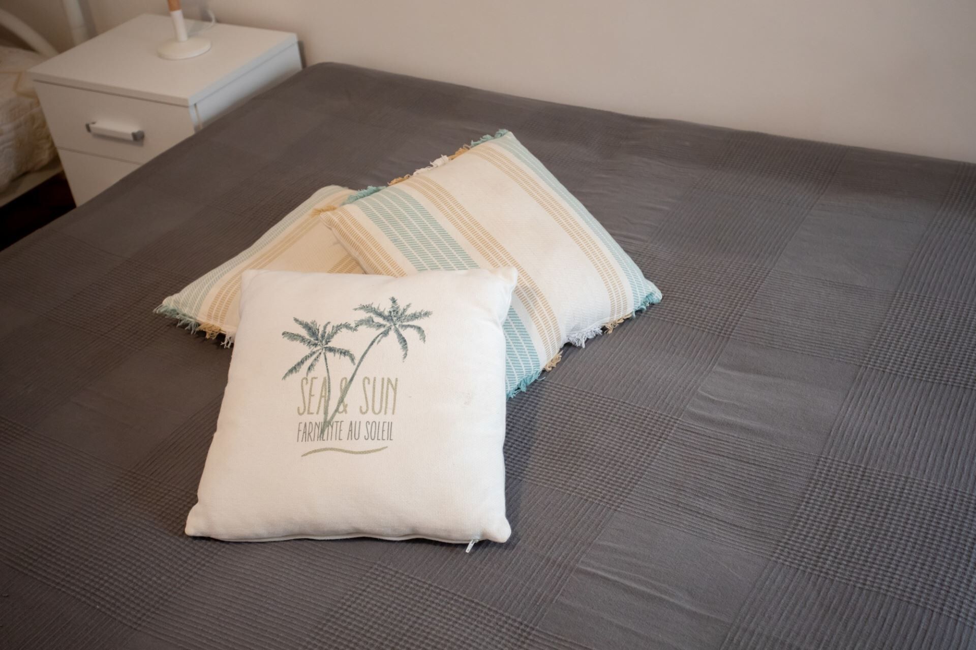 A bed with a dark gray mattress, a white pillow with a palm tree design, and a striped pillow.