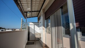 Un balcón cubierto con barandilla de metal, una ventana con persiana decorativa y un cielo azul despejado al fondo.