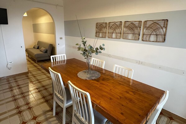 A dining area with a wooden table, white chairs, and a vase with green foliage. There is a sofa and a wall-mounted TV in the background.