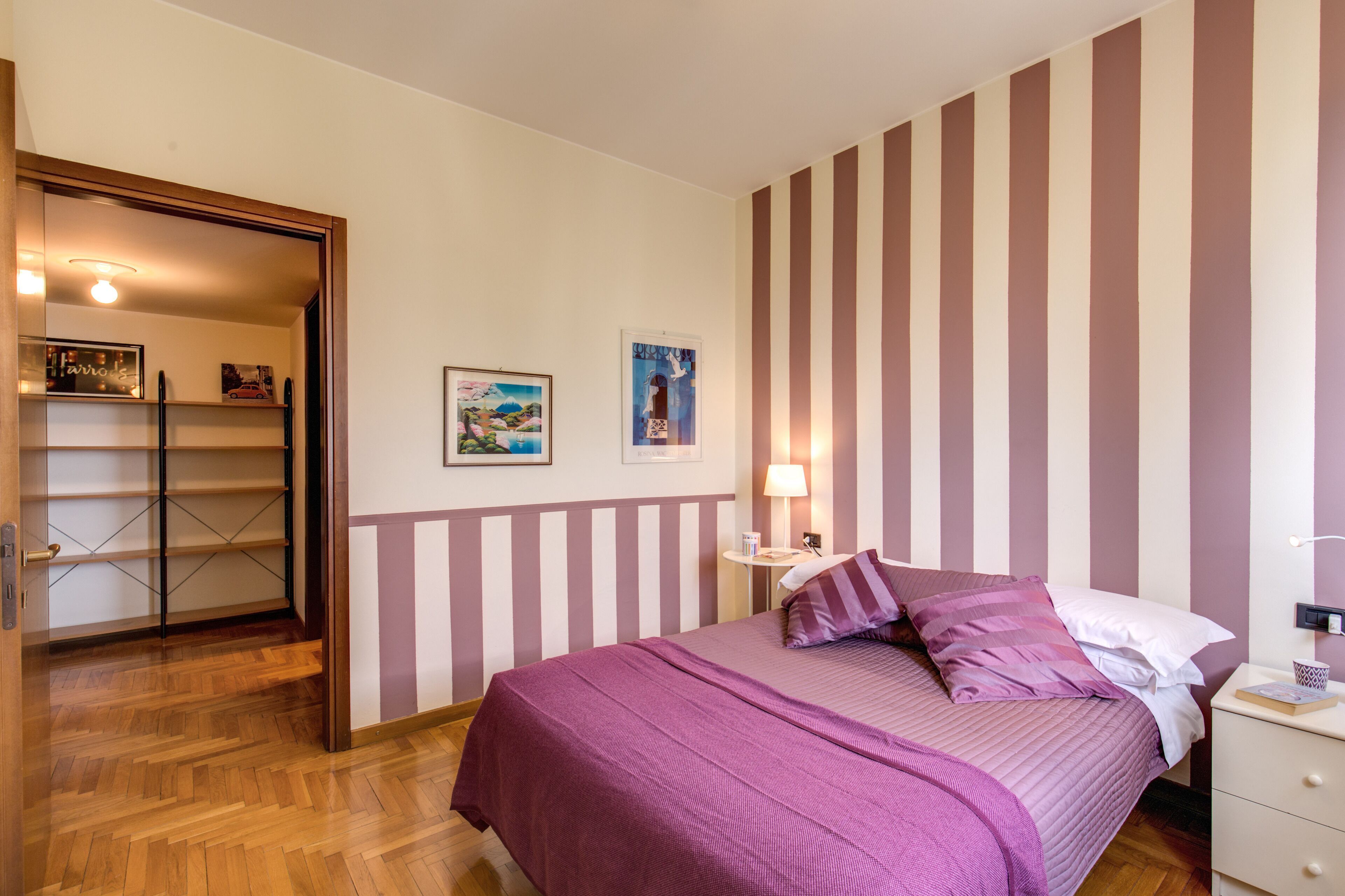 Een slaapkamer met een bed, nachtkastje, lamp en een plank met boeken.