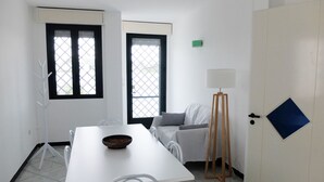 Ein Zimmer mit einem weißen Tisch, zwei Stühlen, einem kleinen Hocker, einem Gemälde auf Staffelei und einer Tür mit einem blauen, rautenförmigen Fenster.