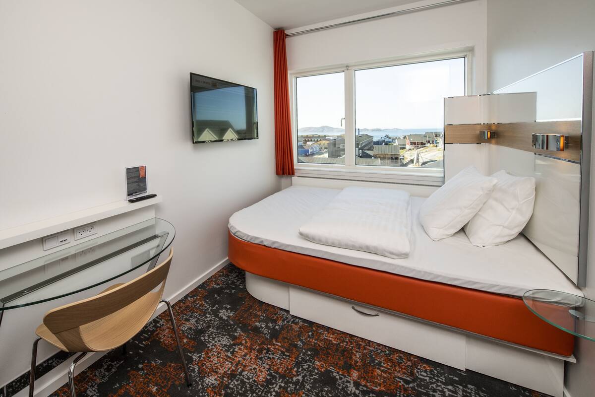A modern hotel room with a bed, a desk, a chair, a TV, and a window with a city view.