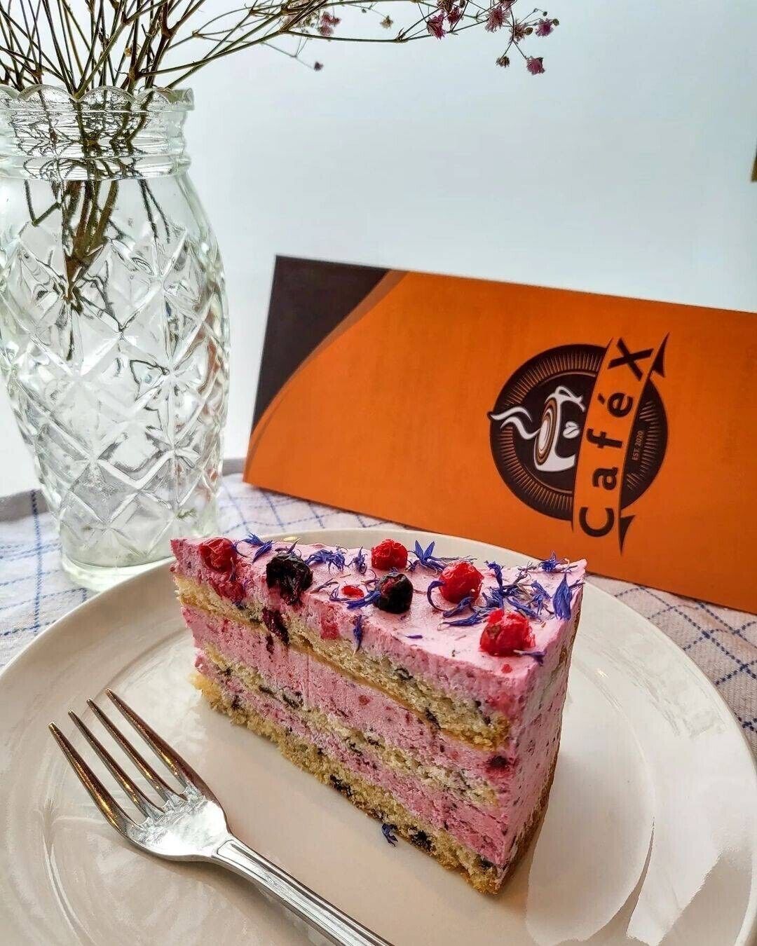 A slice of layered cake with berries on a plate, a fork, and a vase with dried flowers in the background.