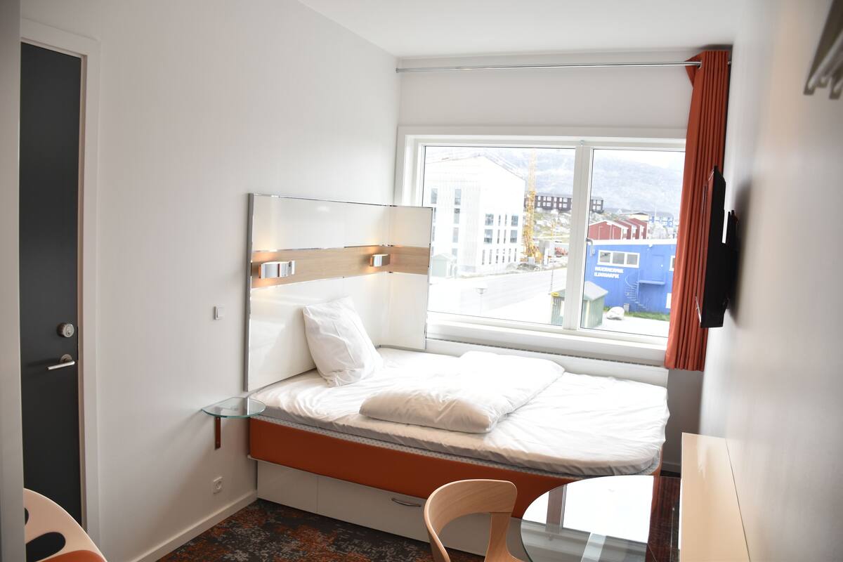 A modern hotel room with a bed, a glass bedside table, a chair, and a window with a view of buildings outside.