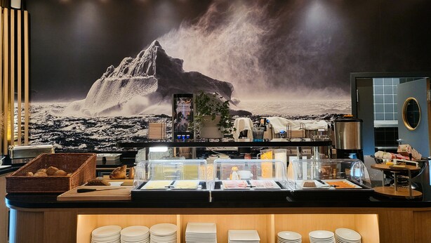 A buffet area with a mountain-themed mural, a fruit bar, and neatly stacked plates. - HHE Express Hotel (Nuuk)