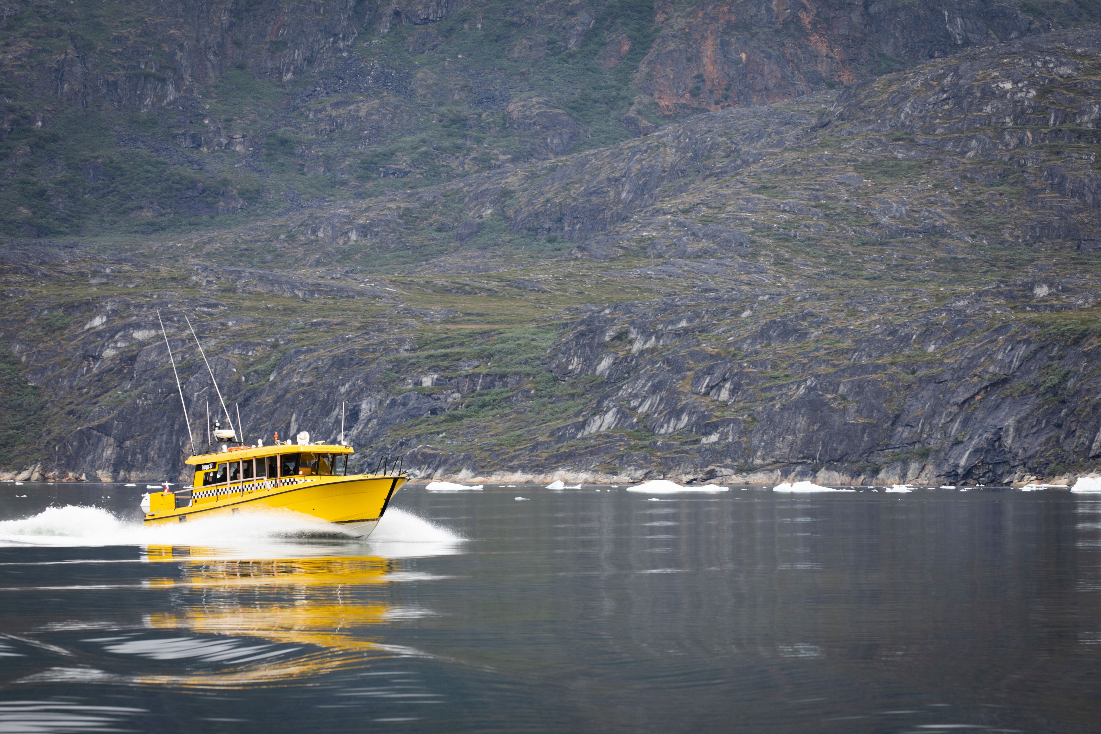 A yellow boat moving through calm water with a rocky, green hillside in the background.