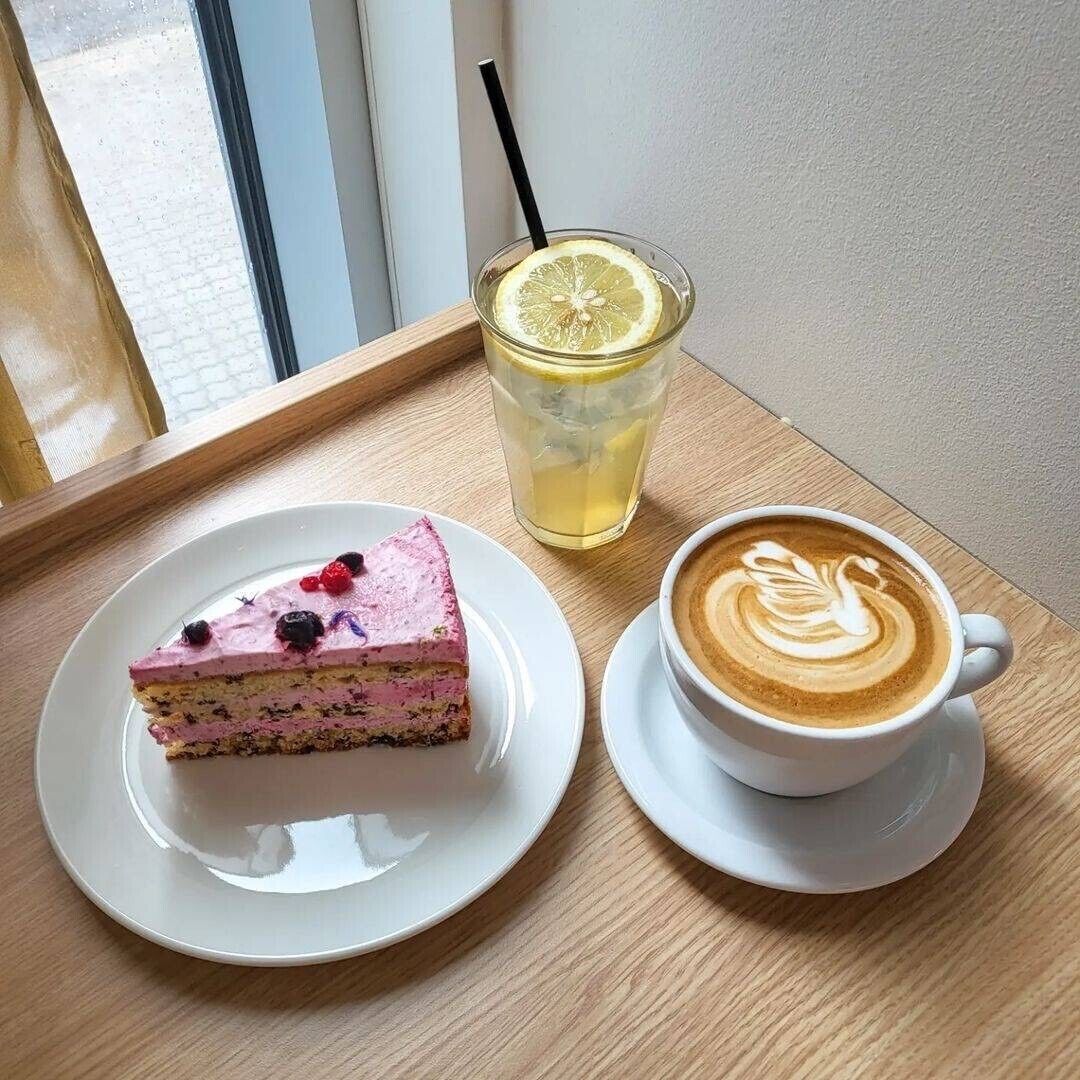A slice of cake with berries on a plate, a glass of lemonade with a lemon slice and straw, and a cup of coffee with latte art on a table.
