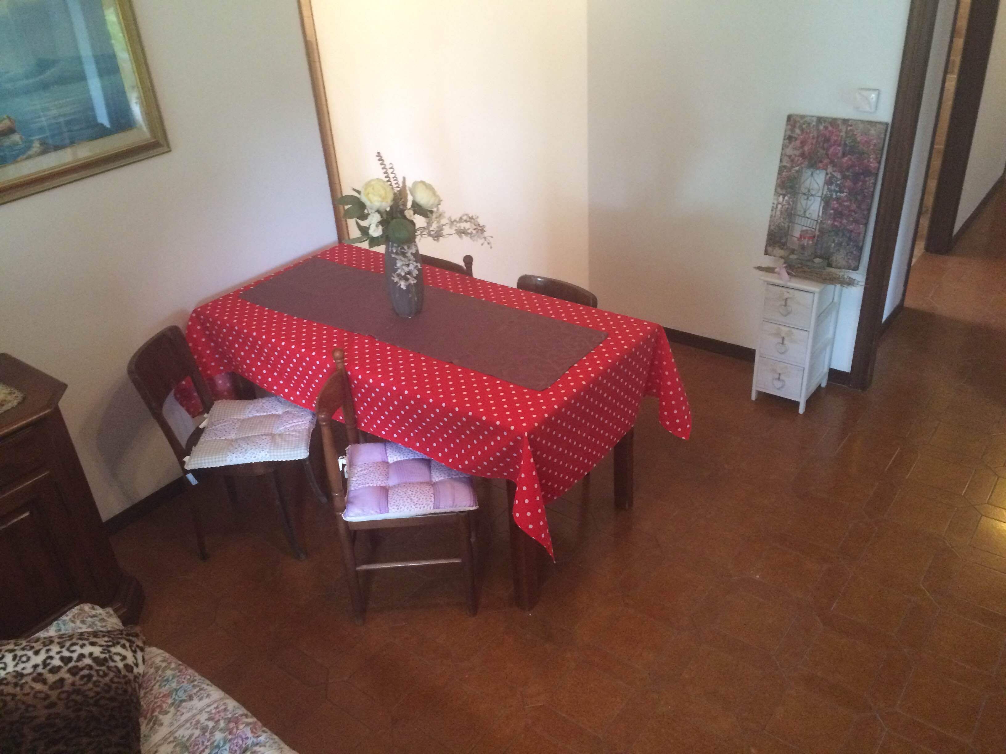 A dining area with a red tablecloth, wooden chairs, and a vase with flowers.