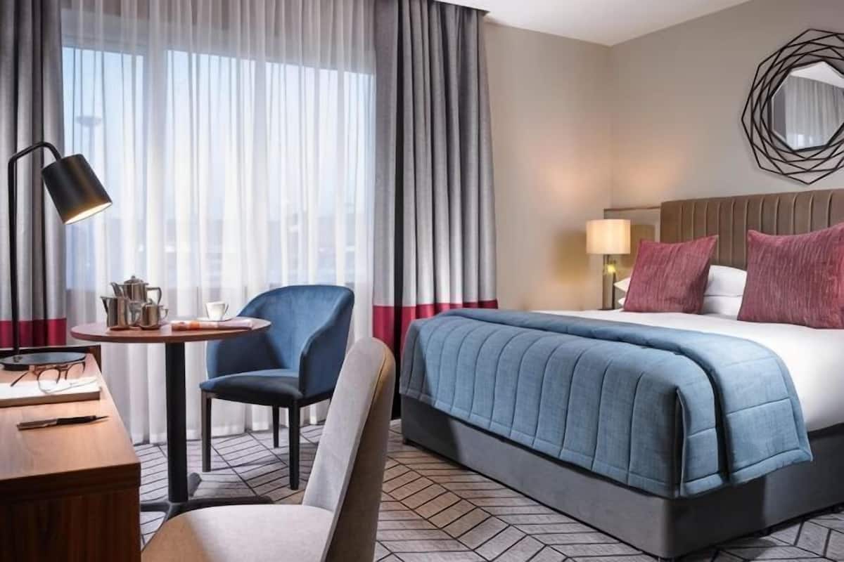 A hotel room with a bed, a desk with a lamp, a chair, a round table with a lamp, and a view through sheer curtains.