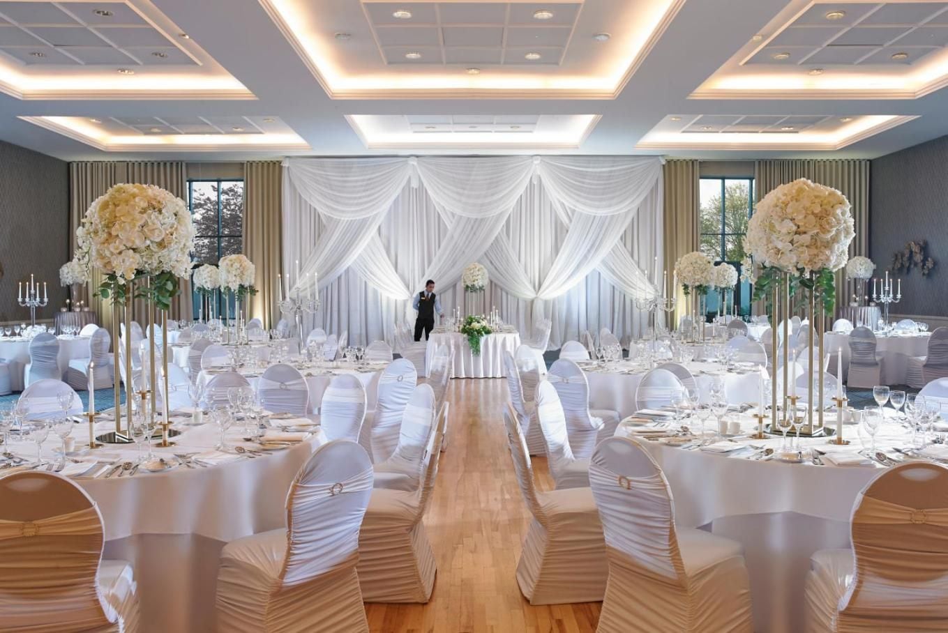 Un salón de banquetes con mesas redondas, cortinas blancas y altas composiciones florales.