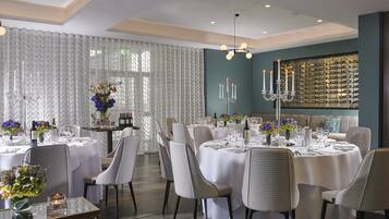 A formal dining room with multiple tables set for a meal, elegant chairs, and a wine rack on the wall.