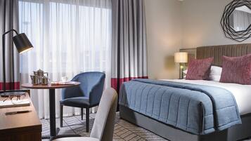 Una habitación de hotel con cama, escritorio, silla, mesa con lámpara y ventana con cortinas.