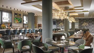 A modern restaurant with a bar area, green upholstered chairs, and a marble-topped dining table.