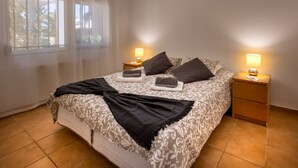 Un lit bien fait, recouvert d’une couvre-lit à motifs, deux tables de chevet avec des lampes et une fenêtre ornée de rideaux légers.