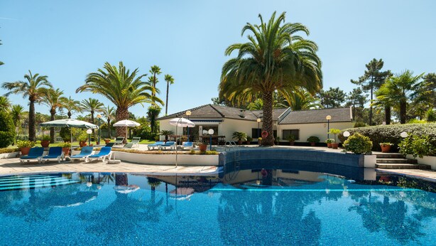 Une piscine entourée de palmiers et d’un bâtiment doté d’un toit en tuiles, avec des chaises longues et des parasols à proximité.