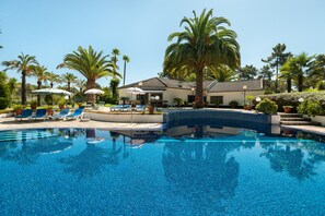 Une piscine entourée de palmiers et d’un bâtiment doté d’un toit en tuiles, avec des chaises longues et des parasols à proximité.