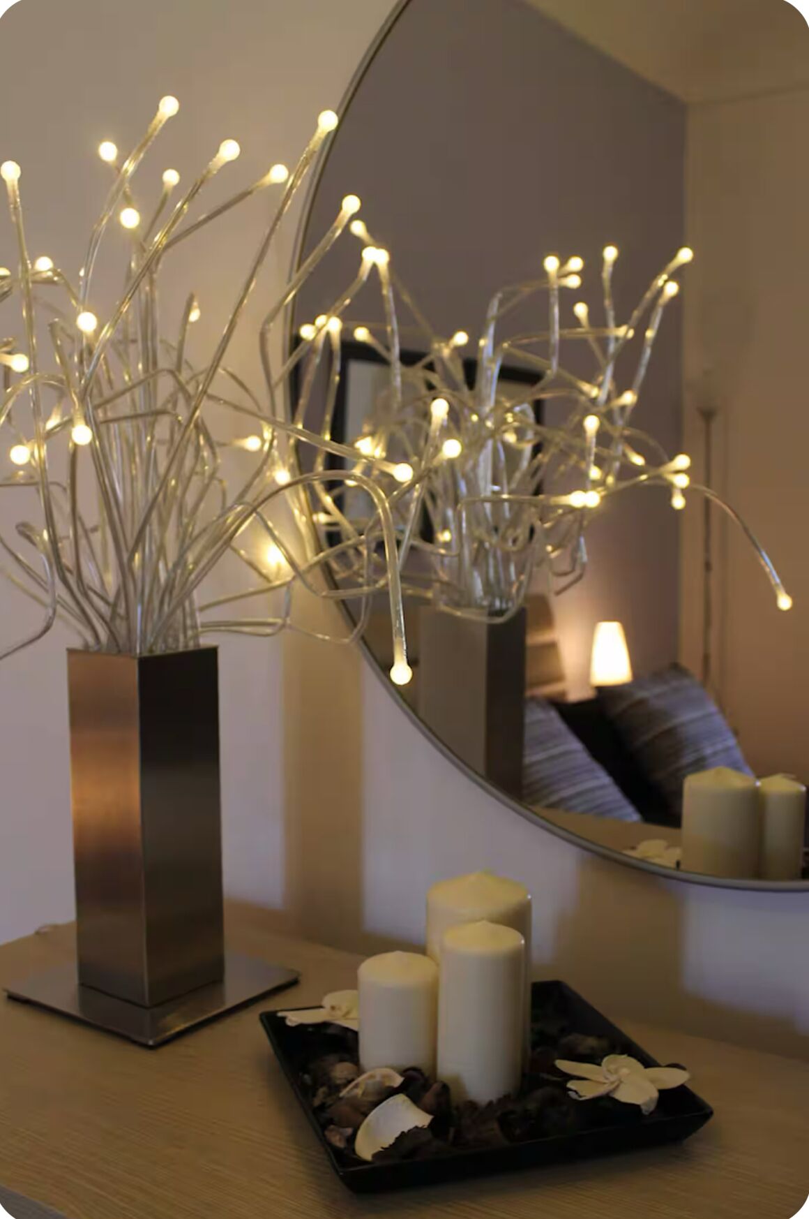 Um interior moderno com um vaso metálico contendo ramos com luzes, um espelho refletindo um ambiente e um tabuleiro com velas e flores.