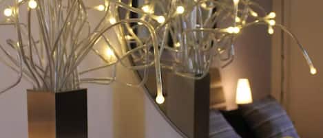 A modern interior with a metallic vase containing branches with lights, a mirror reflecting a room, and a tray with candles and flowers.